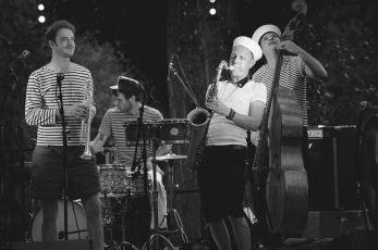 Un groupe de musique jazz sur scène, déguisé en matelot, jouent du saxophone, de la trompette, de la batterie et de la contrebasse.