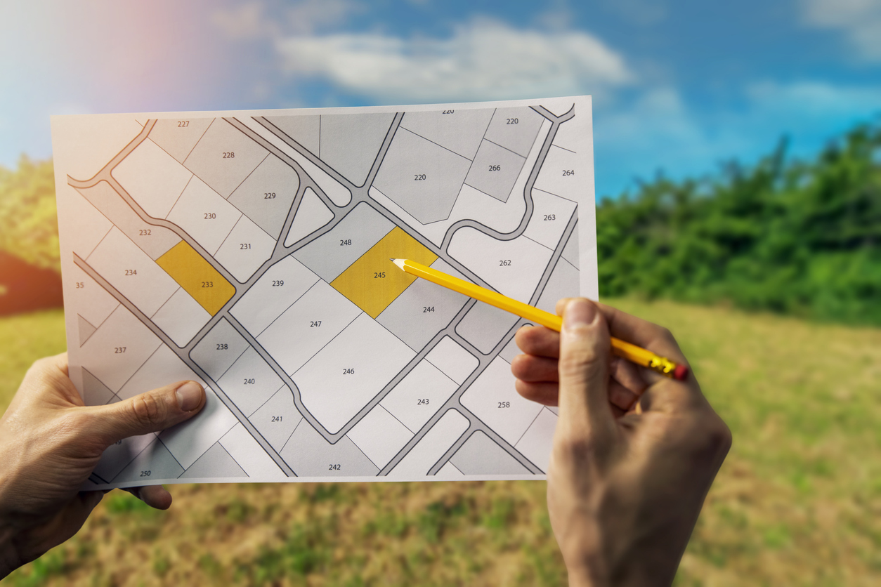 Plan cadastrale sur lequel 2 parcelles sont mises en couleur et pointé du bout d'un crayon par la personne qui tient le plan en l'air, avec en arrière plan un champ